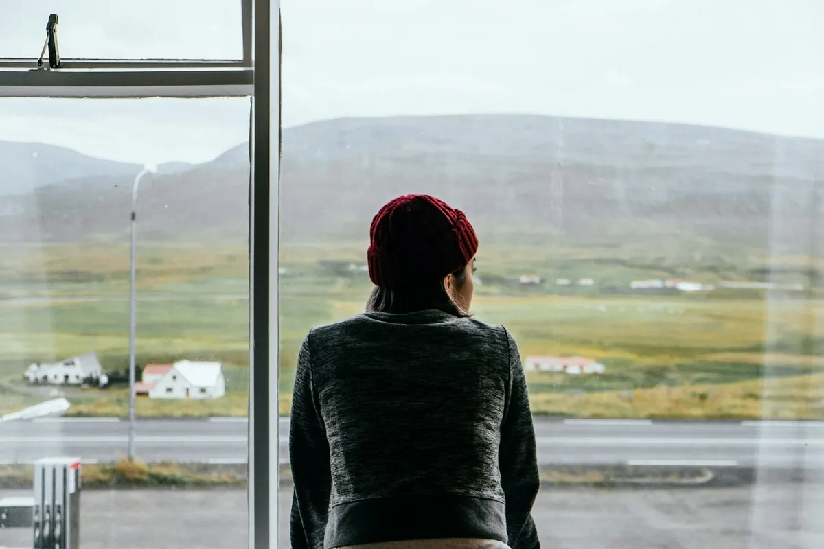 A woman in a red beanie seen from behind, looking out a large window over open green hills