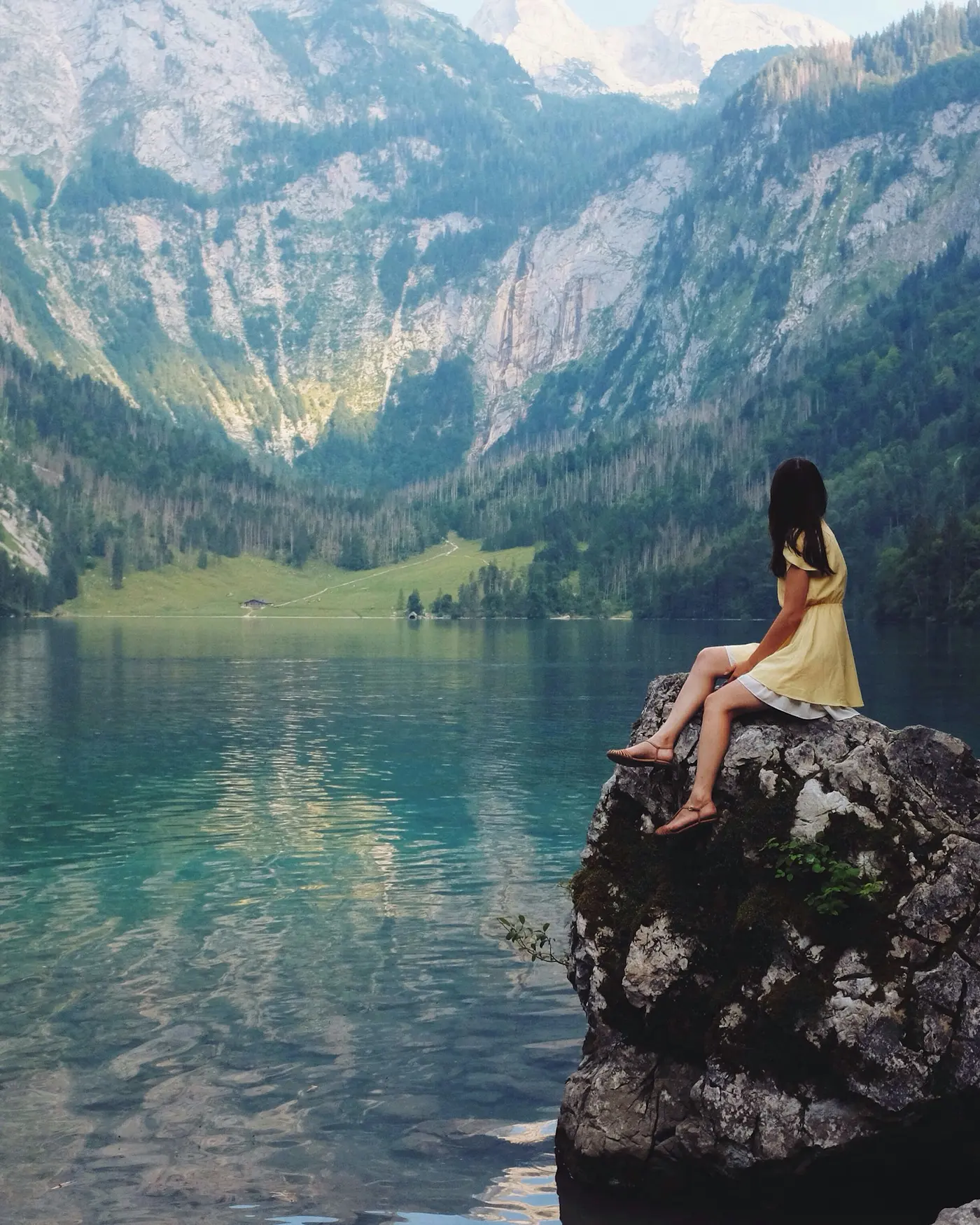A woman travelling solo sits on a rock, looking out over a still alpine lake and mountains