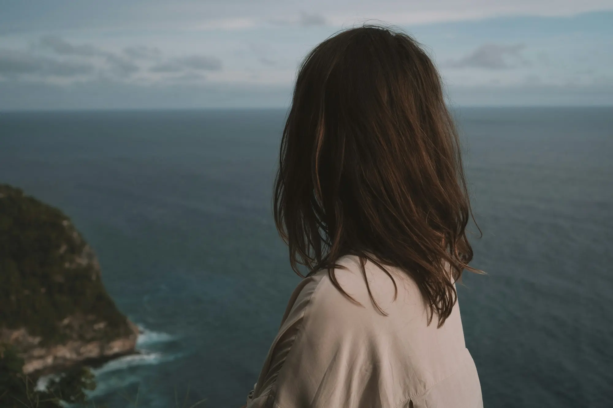 A woman stands alone at a coastal overlook, seen from behind, looking out over the sea