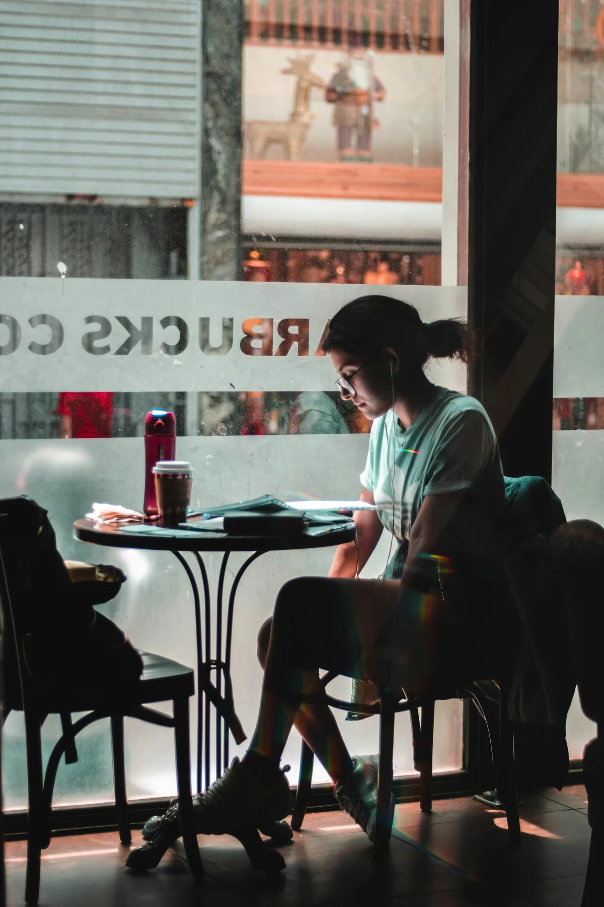 A woman sitting alone by a cafe window with a warm drink, looking out at the street