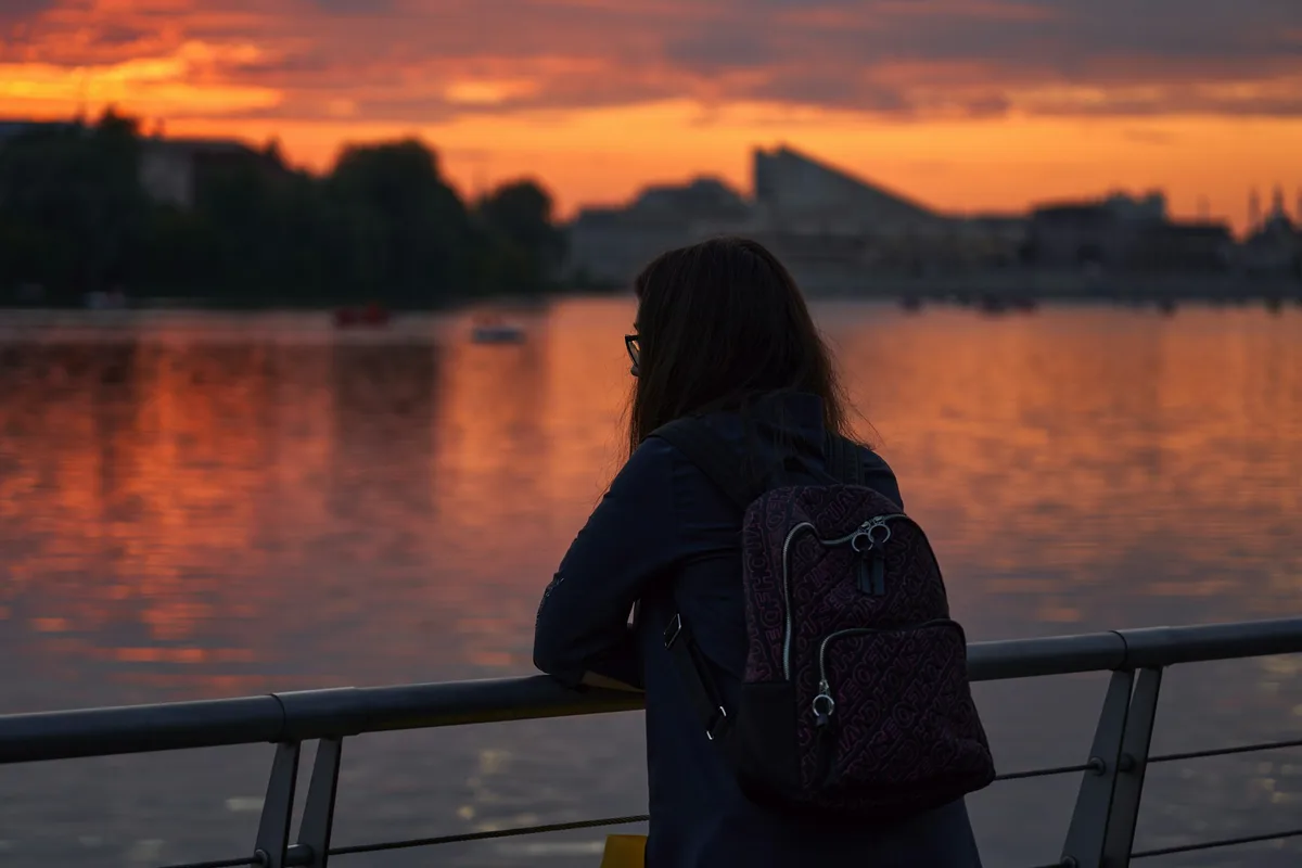 A woman with a backpack standing alone and looking out over a wide body of water
